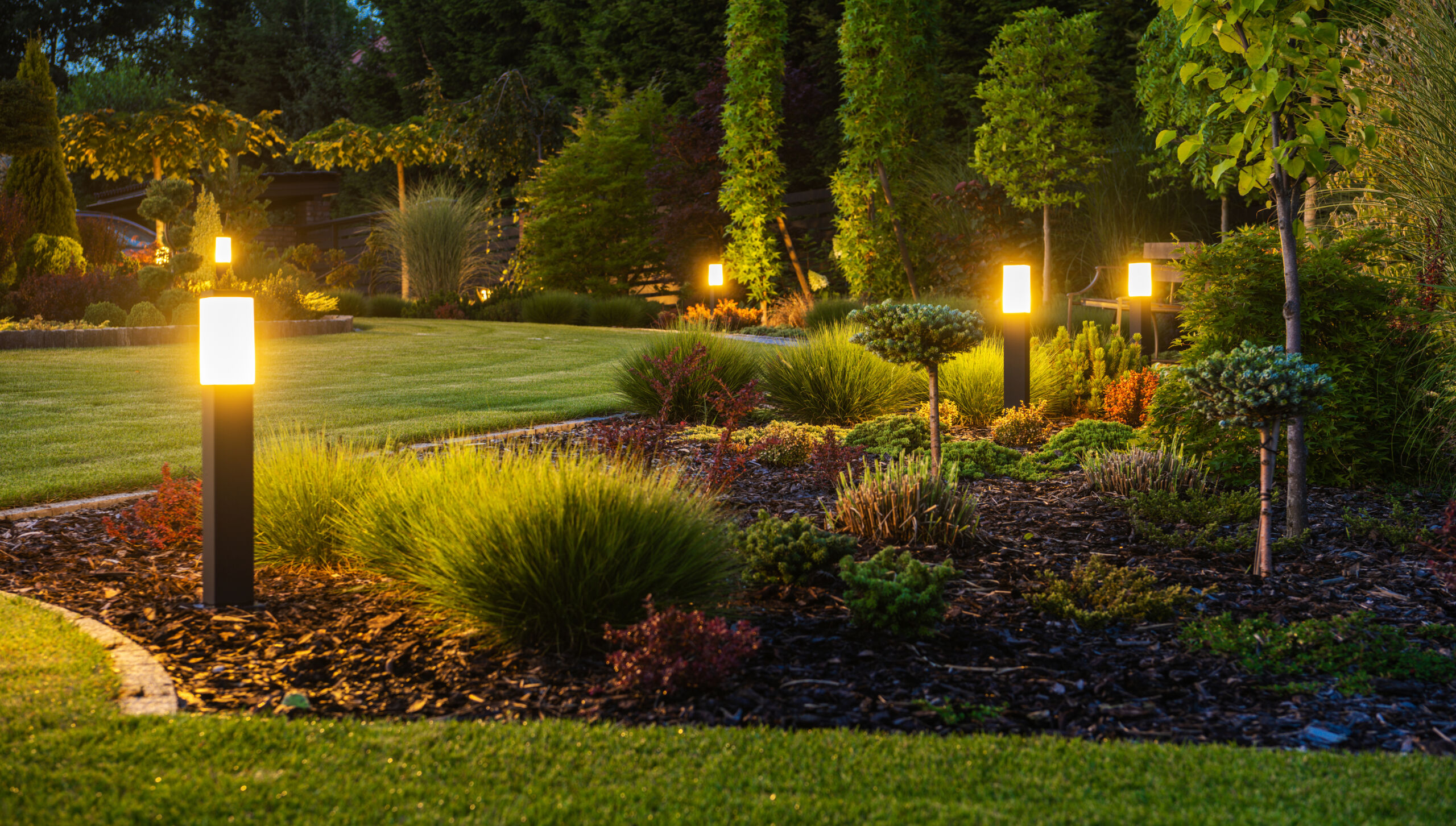 Panoramic Photo of LED Light Posts Illuminated Backyard Garden During Night Hours. Modern Backyard Outdoor Lighting Systems.a